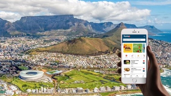 Image of someone holding a phone with the FOMO app open on it superimposed over an image of Table Mountain and the Cape Town Stadium