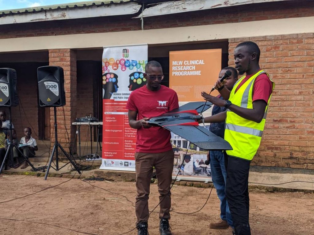 Patrick Kalonde, one of the drones pilots, speaks into a microphone as he briefs the community about the malaria project while his colleague Kennedy Zembere holds a drone in the Kasungu district in Malawi. May 30, 2020. Thomson Reuters Foundation/ Handout via Kennedy Zembere for Malawi Liverpool Wellcome Trust. Photo: Thomson Reuters Foundation/ Charles Pensulo